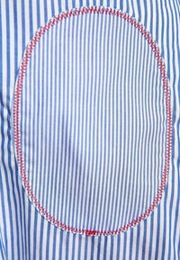 Tela a rayas con líneas verticales azules y blancas, con un parche circular delineado con costuras rojas. Textura suave y diseño a medida.