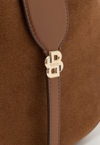 Sac à main en daim marron avec une sangle en cuir lisse et un détail de logo en métal doré. La texture est douce, avec une couture soignée le long des bords.