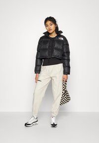 Jachetă puffer neagră, croită, cu guler înalt și logo, asortată cu pantaloni largi crem, adidași negri și albi, și o geantă carouri.