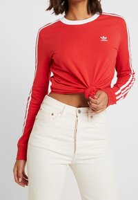 Röd långärmad skjorta med vit krage och tre vita ränder på ärmarna, knuten i midjan. Bärs med ljusa högmidjade jeans.