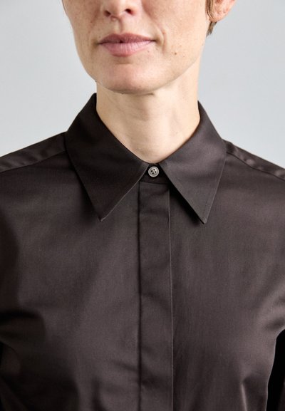 Dettaglio ravvicinato di una persona che indossa una camicia marrone scuro con colletto a punte, mostrando la parte inferiore del viso e il collo.