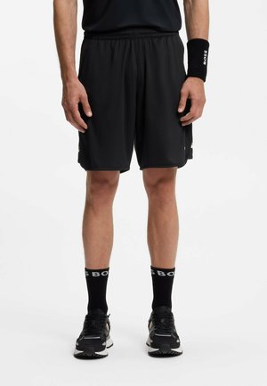 Mann trägt schwarze Sportshorts, schwarzes T-Shirt, schwarzes Armband mit weißem Text, schwarze Socken mit weißem Text und schwarze Laufschuhe.