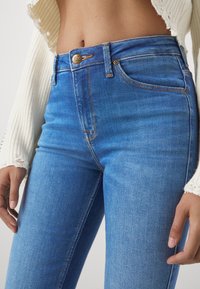 Gros plan de la taille portant un jean Lee bleu taille haute et un pull court blanc aux bords effilochés. Mains détendues sur les côtés.