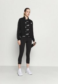 Fekete sportdzseki logó részletekkel, fekete mintás felsővel és fekete leggingssel párosítva. A modell fehér tornacipőt visel, kezében egy vízpalackkal.