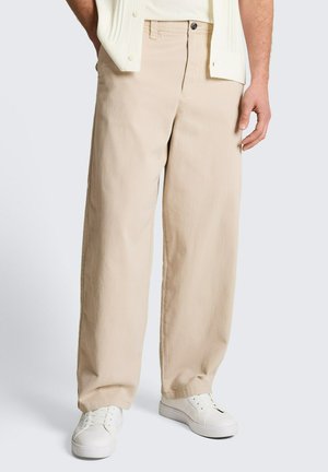 Beige Hosen mit weitem Bein, getragen mit weißen Turnschuhen und einem cremefarbenen Hemd, die Hände teilweise in den Taschen ruhend vor einem einfarbigen Hintergrund.