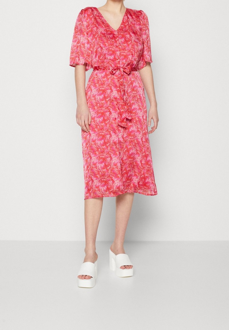 Robe midi florale rose à manches courtes et décolleté en V, avec une taille à nouer ; portée avec des mules blanches à plateforme. Tissu lisse et léger.