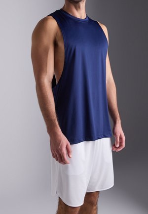 Mann trägt ein ärmelloses dunkelblaues sportliches Tanktop und weiße Sportshorts vor einem einfarbigen Hintergrund.