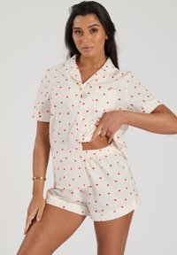 Ensemble pyjama en tissu léger comprenant un haut à manches courtes et un short, avec des motifs de cœurs rouges sur fond crème. Il dispose d'une poche frontale et d'une taille élastique.