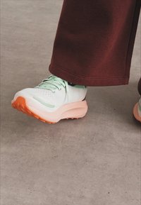 Zapatilla de running ligera en blanco y verde menta con suela naranja, con parte superior de malla, diseño redondeado y cordones planos.