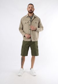 Giacca di jeans beige con due tasche sul petto, pantaloni cargo verde oliva e sneakers bianche con suola testurizzata. Il modello si trova su uno sfondo neutro.