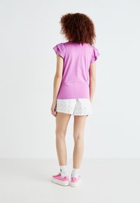 Lilla kortermet topp med volanger, kombinert med hvite broderte shorts. Modellen har på seg rosa sneakers og hvite ankelstrømper.