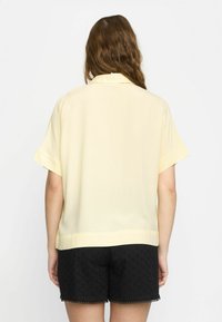 Short-sleeve, collared blouse in soft yellow fabric. Loose fit with a straight hem, paired with black embroidered shorts featuring a scalloped edge.