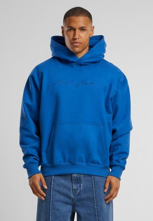 Mėlyna hoodie, pagaminta iš minkštos medžiagos, turi priekinę kišenę ir raukšlėtus rankogalius. Prekės ženklas yra išsiuvinėtas kontrastinga spalva.
