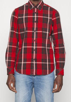 Homme portant une chemise à carreaux rouge, noire et blanche à manches longues et un jean bleu clair, debout devant un fond uni.