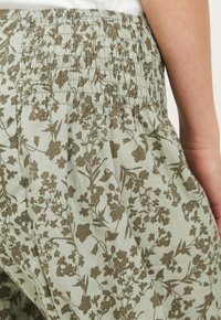 Nærbillede af talje og hofte iført lys grønne bukser med brune blomsterprint og elastisk smock ved taljen.