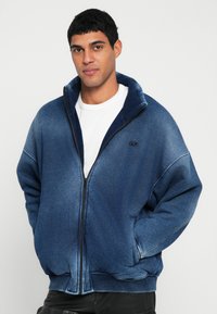 Modra jopa z zadrgo in prelivajočo teksturo. Ima stoječi ovratnik, rebraste manšete ter logotip na levi strani prsnega koša. Prijeten, sproščen kroj.