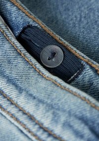 Denimjeans i lys blå farge, med kontrasterende brune sømmer og en plastknapp med et mørkeblått stoffbånd på innsiden av linningen.