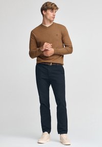 Brun v-ringad tröja, mörka byxor och beige skor. Tröjan har en slät textur med en liten logotyp på bröstet. Avslappnad passform.