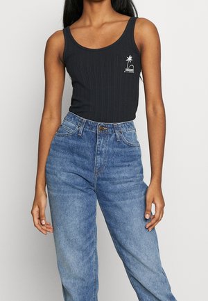 Femme portant un débardeur noir sans manches côtelé avec un petit logo palmier blanc et un jean bleu taille haute sur un fond uni.