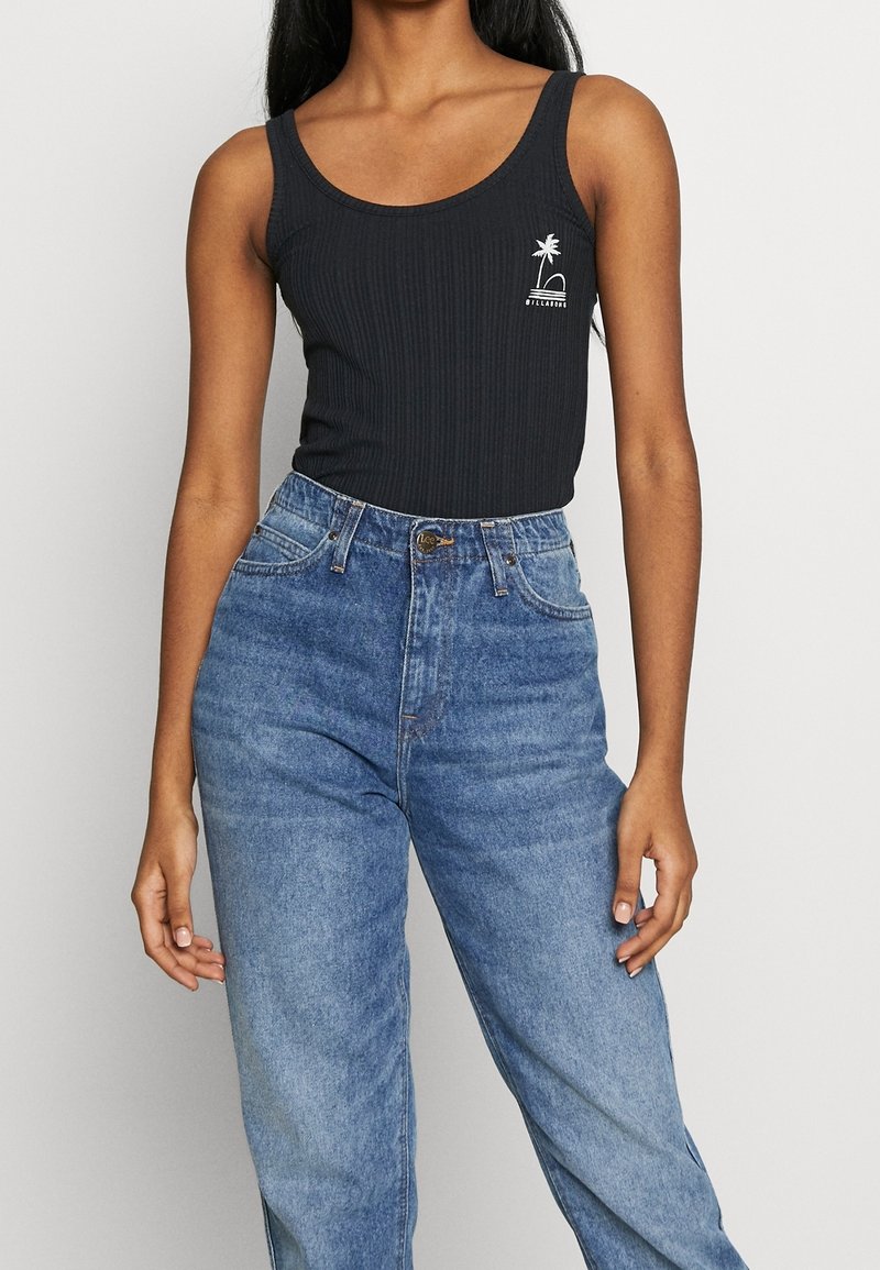 Femme portant un débardeur noir sans manches côtelé avec un petit logo palmier blanc et un jean bleu taille haute sur un fond uni.
