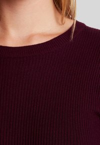 Burgunderfarbene, gerippte Strickoberteil mit Rundhalsausschnitt und strukturiertem Stoff. Nahaufnahme zeigt feine Details und ein einheitliches Muster.