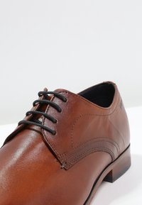 Chaussure habillée en cuir marron avec lacets noirs, coutures détaillées, et un petit talon empilé sur une surface blanche.