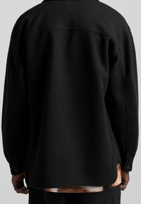 Černá vlněná košile s bundou, která má límec, knoflíkové manžety a mírně zakřivený lem. Má jemnou texturu a minimalistický design.