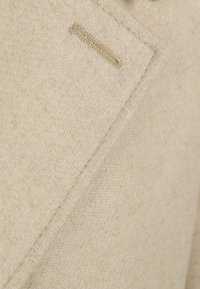 Beige wollen blazer met een visgraatpatroon, scherpe revers en subtiele stikdetails. Gelaagd stof met een zachte afwerking.
