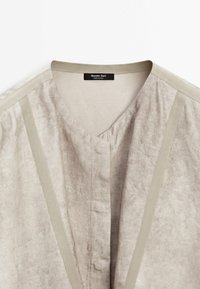 Haut en lin gris clair avec un col en V, présentant des accents beiges contrastants et un design minimaliste. Comprend une étiquette au niveau de l'encolure.