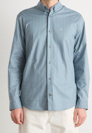 Mann trägt ein hellblaues Hemd mit weißen vertikalen Streifen und weißen Hosen, stehend vor einem einfarbigen Hintergrund.