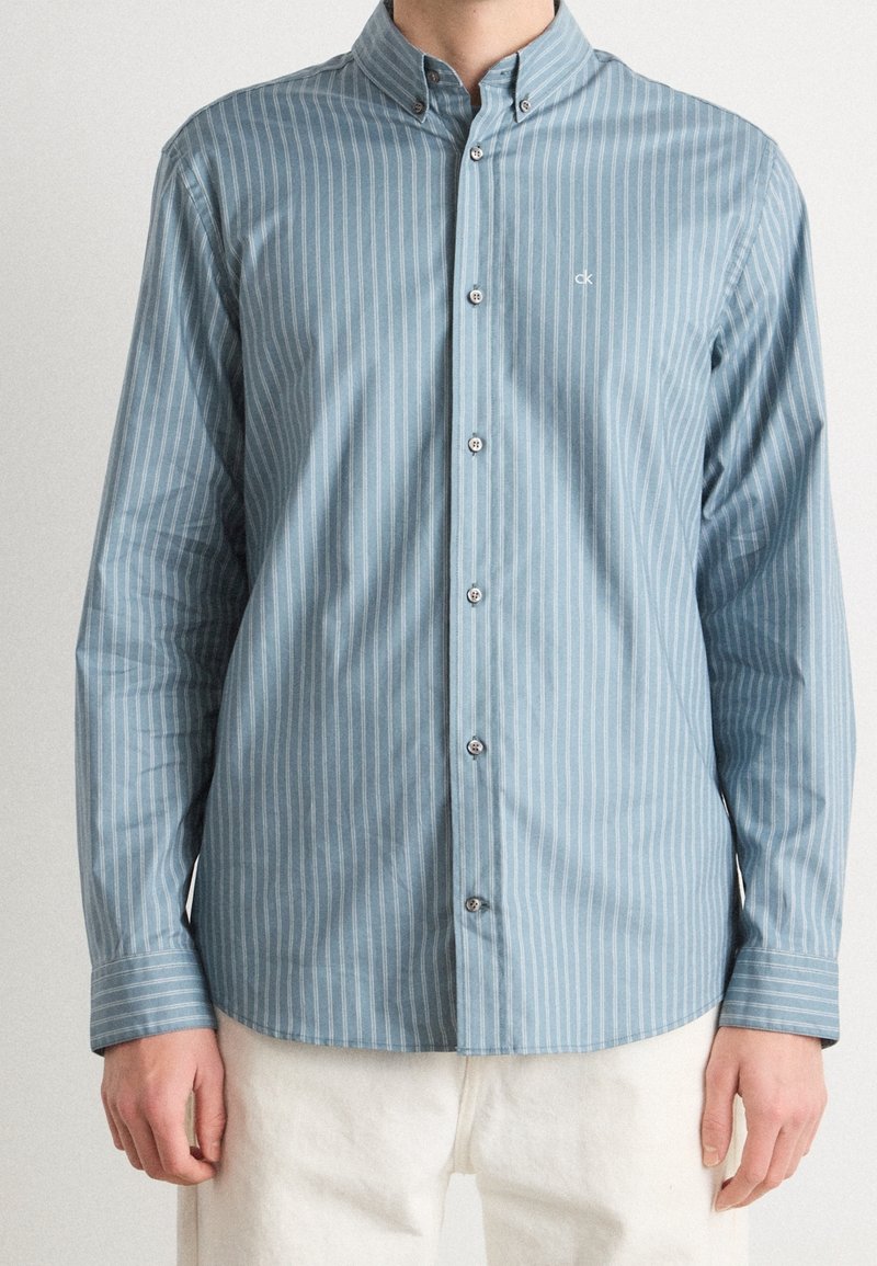 Homme portant une chemise bleu clair boutonnée avec des rayures verticales blanches et un pantalon blanc, debout devant un fond uni.