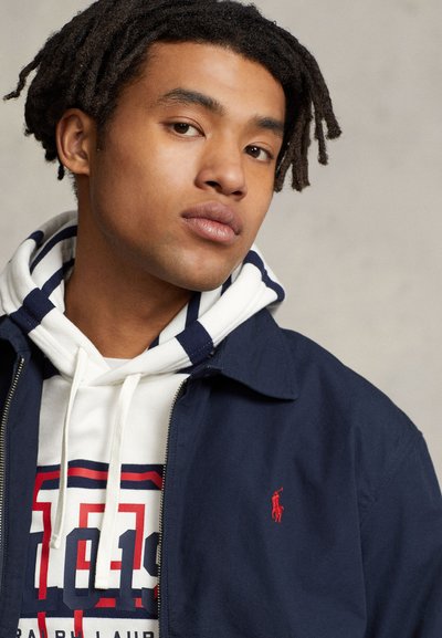 Young man with dreadlocks wearing a navy jacket and striped hoodie, gazing slightly to the side against a neutral background.