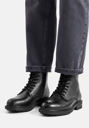 Czarne botki z skóry o fakturowanym wykończeniu, zaokrąglonym nosku, sznurowane, z wytrzymałą podeszwą. Zaprezentowane z ciemnymi, podwiniętymi spodniami dżinsowymi.
