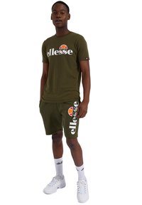 Alie žalios spalvos marškinėliai su "ellesse" logotipu, derinti su atitinkamais šortais. Baltos akcentai ir logotipo detalės; baltos sportbačių ir kojinių derinys užbaigia įvaizdį.