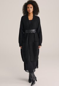 Svart stickad cardigan med långa ärmar, två framfickor och bälte. Buren över en plisserad svart klänning och högklacksstövlar.