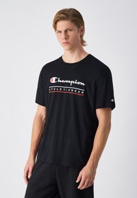 Camiseta de algodón negra con cuello redondo, que presenta un logo blanco y acentos rojos. Manga corta y corte relajado. Diseño simple sin adornos.