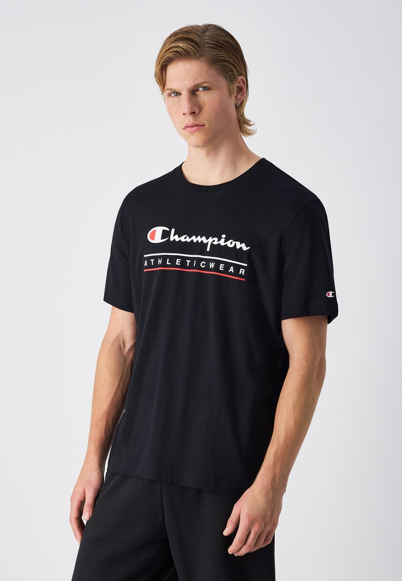 Camiseta de algodón negra con cuello redondo, que presenta un logo blanco y acentos rojos. Manga corta y corte relajado. Diseño simple sin adornos.