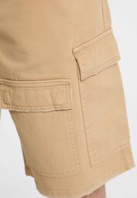Shorts cargo beiges en mélange de coton, avec deux poches latérales, ourlet effiloché et détails de couture visibles. Design décontracté.