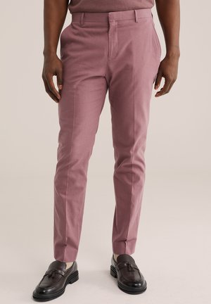 Moški, oblečen v ozke vijolične hlače z žepoma spredaj, obut v temno rjave usnjene loaferje z cofki in bež nogavice na enobarvnem ozadju.