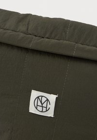 Κοντινό πλάνο από σκούρο πράσινο καπιτονέ ύφασμα με μια λευκή ορθογώνια ετικέτα που έχει ένα μαύρο κυκλικό αφηρημένο λογότυπο.