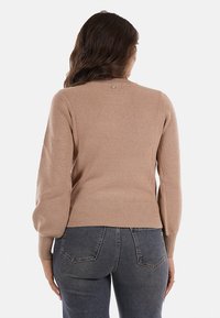 Un maglione beige chiaro con texture a coste, maniche lunghe e orlo aderente. Presenta un piccolo dettaglio metallico sul retro del collo.