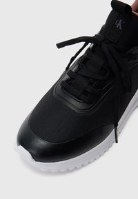 Μαύρο αθλητικό παπούτσι με συνδυασμό υφασμάτινης υφής και λείας δερματίνης, με λευκή καουτσούκ σόλα και επίπεδα κορδόνια. Έντονη λεπτομέρεια λογότυπου παρούσα.