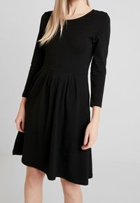 Svart långärmad klänning i mjukt material, med rund halsringning, figursydd överdel och utsvängd kjol med veck som börjar vid midjan.