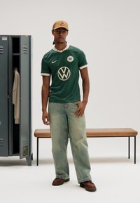 Zöld sportmez fehér díszítésekkel és Volkswagen logóval, világoskék farmerrel és barna cipővel párosítva. Szürke öltözőszekrény és fapad a háttérben.