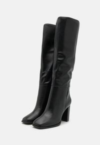 Bottes en cuir noir montant jusqu'au genou, avec une texture lisse, un bout carré et un talon bloc. Caractérisées par une coupe ajustée sans éléments visibles ni ornements.