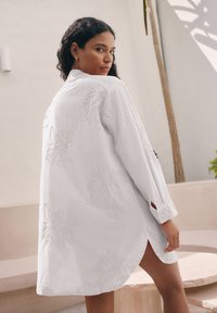 Robe chemise blanche brodée avec des motifs floraux, manches longues avec des accents de boutons et fentes latérales pour un mouvement facile.