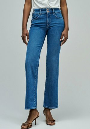 Persona que lleva jeans rectos de tiro alto azules con cierre de doble botón y sandalias marrones de tacón, de pie frente a un fondo liso.