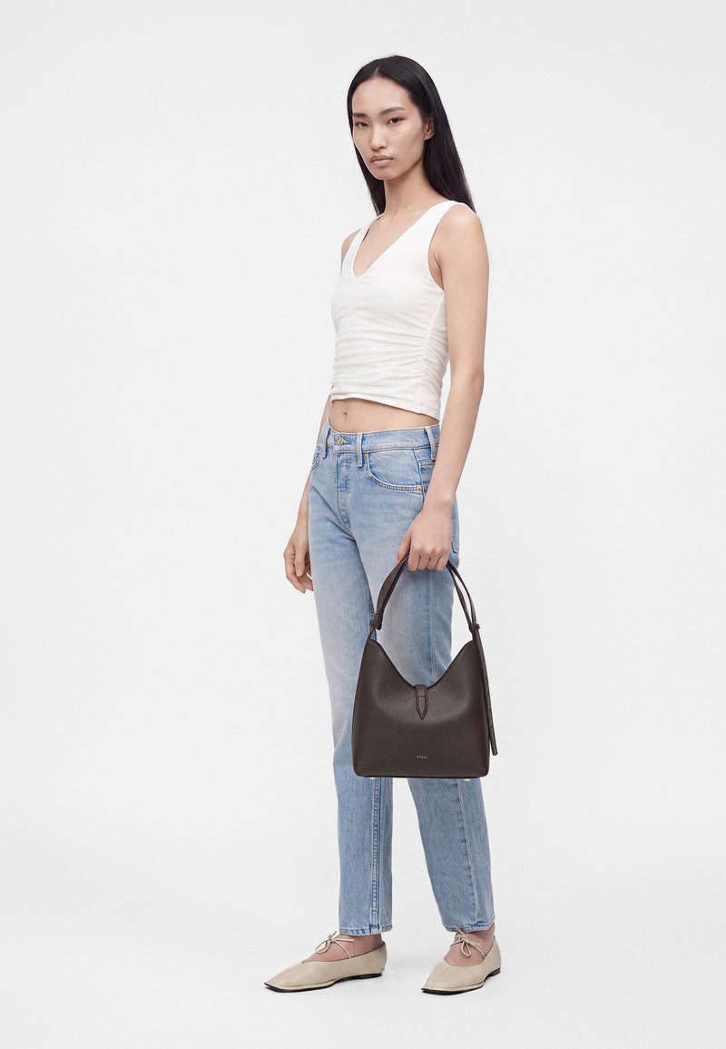 Femme aux longs cheveux foncés portant un top blanc sans manches, un jean bleu clair, des ballerines beige, tenant un sac à main en cuir marron foncé.