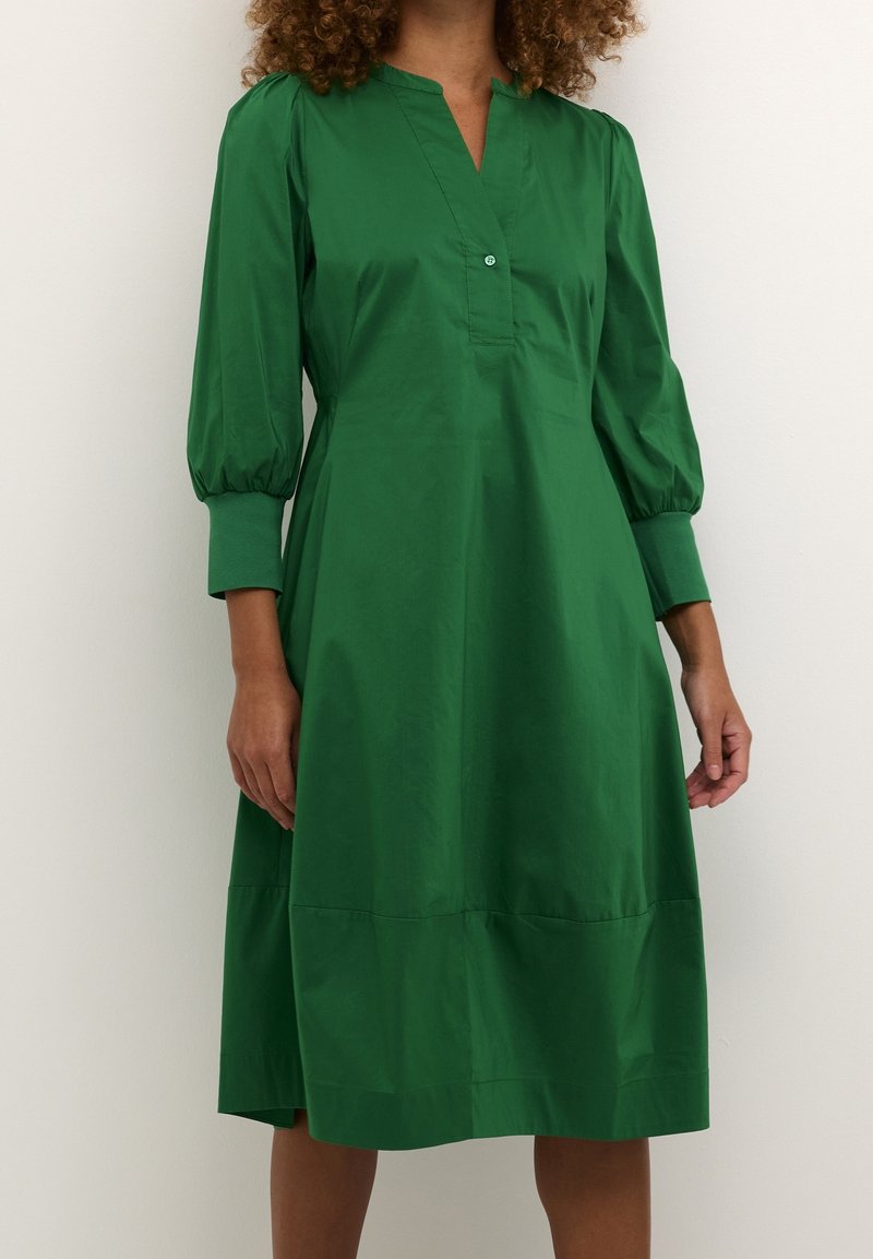 Grön, knälång klänning i lätt tyg. Har krage, knappdetalj och puffärmar. Slät textur.