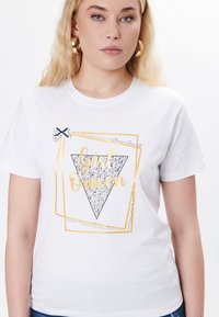 Hvid t-shirt med et trianer grafik, der har "Girl Queen" i guld, glitrende detaljer og perle accenter, suppleret med en sløjfedetalje.
